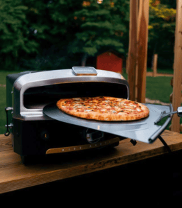 Pizza oven with a pizza cooking outdoors on a wooden table.