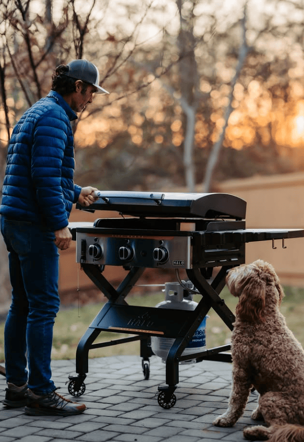 Outdoor grill with a man preparing food, dog sitting nearby, during sunset.