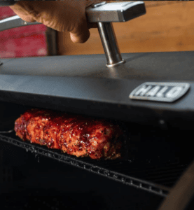 A hand adjusting the temperature on the Halo outdoor smoker with ribs cooking inside.